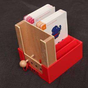 Könnte beinhalten: Ein roter und hölzerner Kartenhalter mit einer hölzernen Schiebetür und einer kleinen Holzkugel. Der Halter enthält mehrere Karten mit Illustrationen und Text. Der Name "EMIL" ist auf der Seite aufgedruckt.