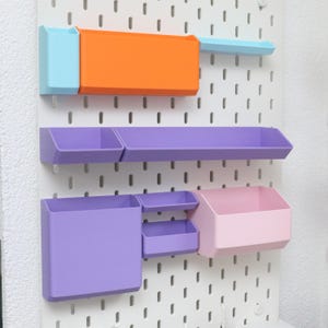 Könnte beinhalten: Eine weiße Lochwand mit mehreren farbigen Aufbewahrungsbehältern. Die Behälter sind in den Farbtönen Orange, Blau, Lila und Rosa gehalten. Die Lochwand hat gleichmäßig verteilte Löcher zur Befestigung der Behälter.