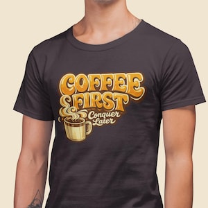 以下が含まれることがあります： オレンジと黄色で「COFFEE FIRST Conquer Later」と書かれた濃い灰色のTシャツ。テキストの下には、湯気の立つコーヒーカップのグラフィックがあります。シャツはクルーネックです。