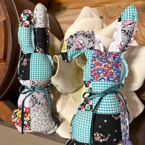 K&ouml;nnte beinhalten: Zwei handgefertigte Patchwork-Stoff-Hasenfiguren. Die Hasen bestehen aus verschiedenen gemusterten Stoffen in Farben wie Schwarz, T&uuml;rkis, Rosa und Blumenmustern. Jeder Hase hat lange Ohren und ist mit einem t&uuml;rkisfarbenen Band um den Hals gebunden.