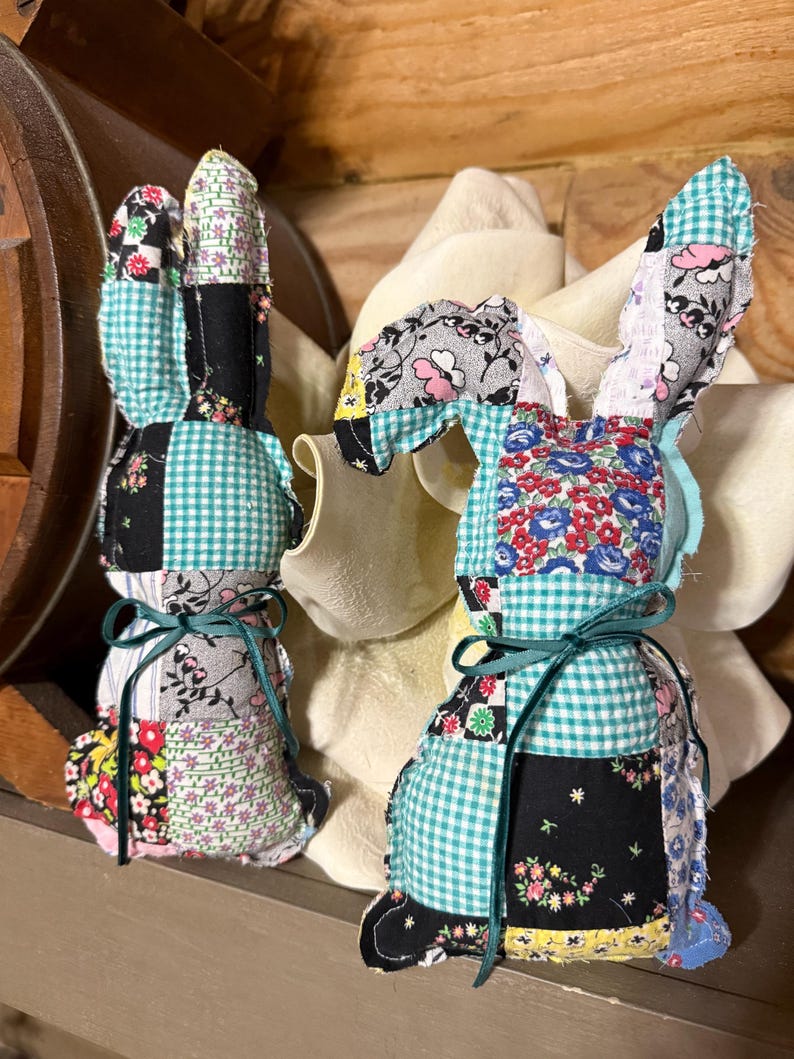 K&ouml;nnte beinhalten: Zwei handgefertigte Patchwork-Stoff-Hasenfiguren. Die Hasen bestehen aus verschiedenen gemusterten Stoffen in Farben wie Schwarz, T&uuml;rkis, Rosa und Blumenmustern. Jeder Hase hat lange Ohren und ist mit einem t&uuml;rkisfarbenen Band um den Hals gebunden.