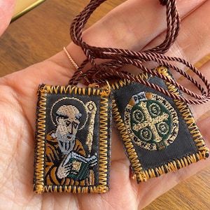 May include: Two rectangular fabric scapulars with brown braided cord. One features an embroidered figure of a bearded person holding a book, while the other displays a green and gold cross emblem. Both have black fabric bases with gold stitching.