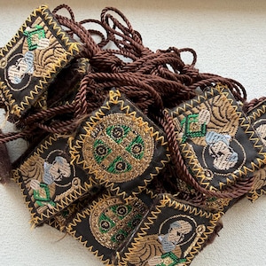 May include: A collection of small, square embroidered patches with religious imagery. The patches are primarily black with gold, green, and blue accents. Each patch is bordered with gold stitching and attached to a brown rope.