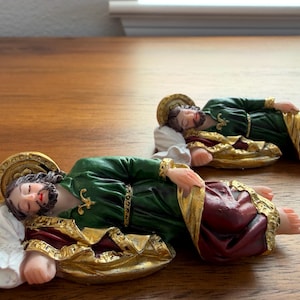 May include: Two identical figurines of a sleeping figure with a beard, wearing a dark green tunic with gold trim and a deep red cloak. Each figure has a gold halo behind the head and rests their head on a white pillow, lying in a horizontal position.