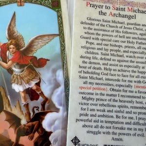 May include: A rectangular prayer card featuring an illustration of Saint Michael the Archangel defeating a demon on the left and the text of the Prayer to Saint Michael the Archangel on the right, printed in black ink on a cream-colored background.