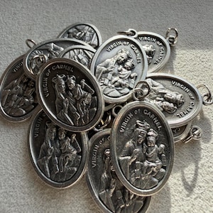 May include: A collection of silver-colored oval pendants. Each pendant features an embossed image of the Virgin of Carmel with the text "VIRGIN OF CARMEL" around the edge. The pendants are linked with small loops.