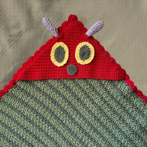 May include: A handmade crochet blanket featuring a red caterpillar head with yellow-rimmed eyes, grey nose, and purple antennae. The blanket's body is green and grey striped. The edges are scalloped.