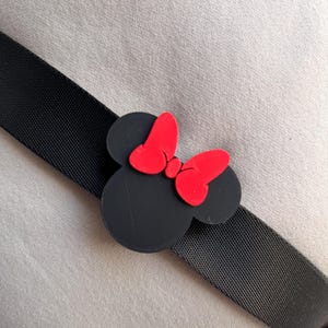 May include: Black Minnie Mouse-shaped accessory with a red bow, attached to a black strap. The accessory is a cartoon-style design, with a solid black head and red bow. The strap is a woven, textured material.