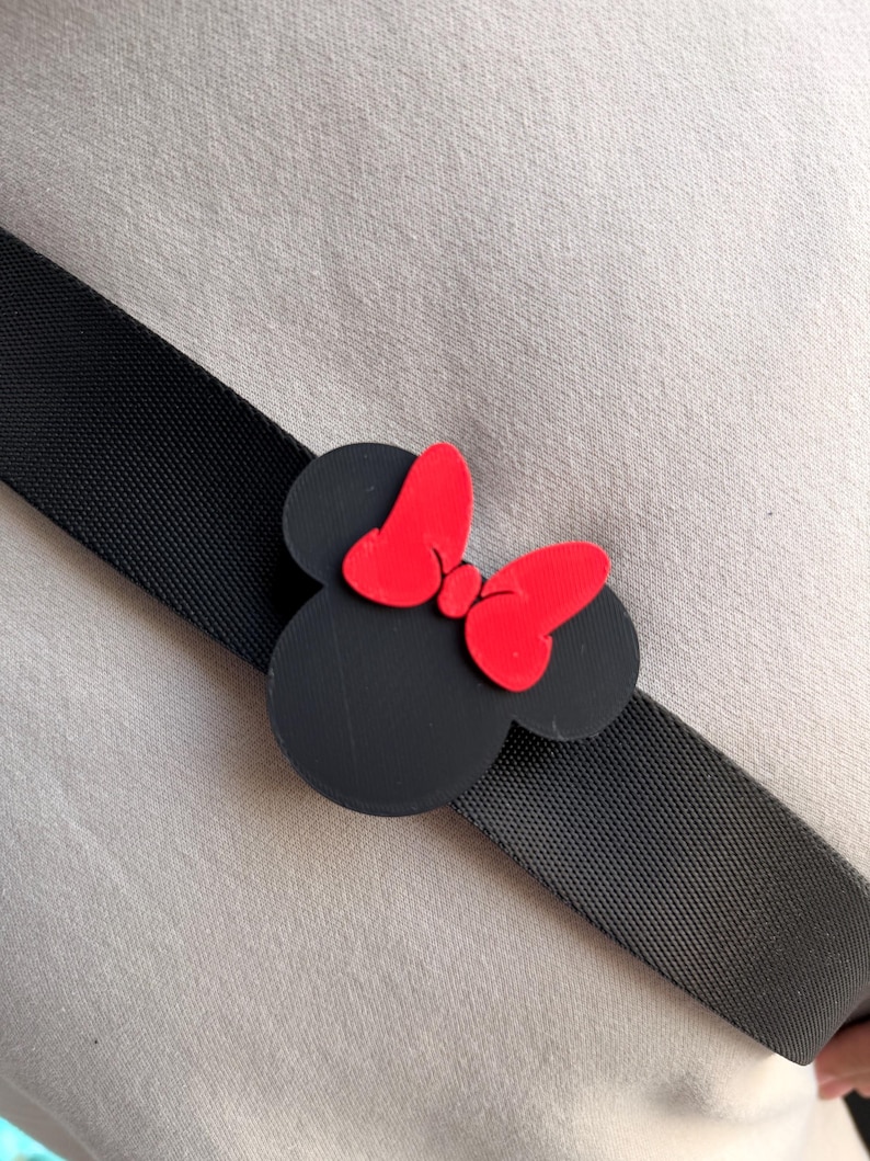May include: Black Minnie Mouse-shaped accessory with a red bow, attached to a black strap. The accessory is a cartoon-style design, with a solid black head and red bow. The strap is a woven, textured material.