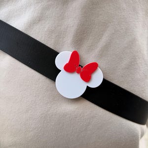 May include: A white Minnie Mouse head-shaped accessory with a red bow is attached to a black strap. The accessory is positioned on a light beige fabric background. The design is a playful nod to a beloved cartoon character.
