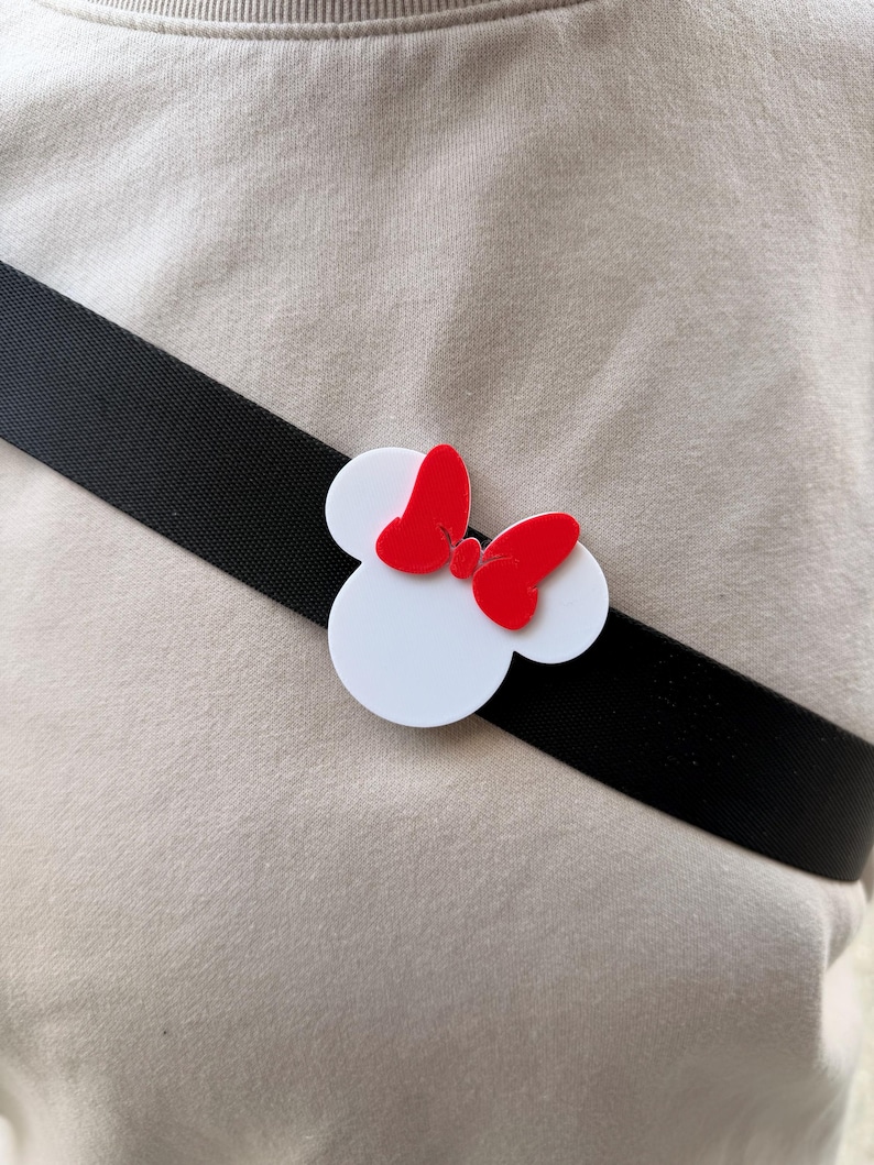 May include: A white Minnie Mouse head-shaped accessory with a red bow is attached to a black strap. The accessory is positioned on a light beige fabric background. The design is a playful nod to a beloved cartoon character.