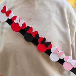 May include: A black strap adorned with colorful Minnie Mouse-shaped embellishments. The embellishments are in various colors, including red, white, black, and pink, each with a matching bow. The strap is angled across a neutral-colored background.