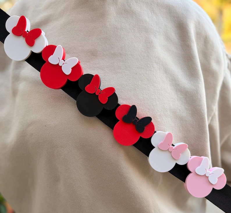 May include: A black strap adorned with colorful Minnie Mouse-shaped embellishments. The embellishments are in various colors, including red, white, black, and pink, each with a matching bow. The strap is angled across a neutral-colored background.