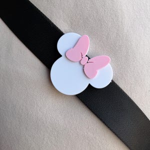 May include: Black elastic band with a white and pink Minnie Mouse bow accent. The bow features a white circular base with a pink bow on top. The band is angled across a neutral background.