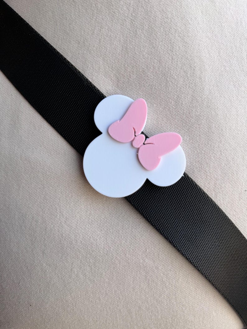 May include: Black elastic band with a white and pink Minnie Mouse bow accent. The bow features a white circular base with a pink bow on top. The band is angled across a neutral background.
