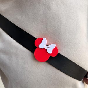 May include: A red and white Minnie Mouse bow pin is attached to a black strap. The pin is a classic design with a red circular base and white bow. The strap is worn across the body over a beige sweatshirt.