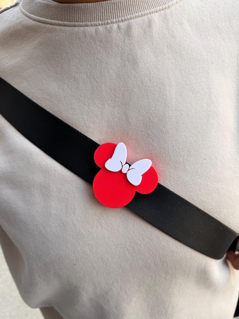May include: A red and white Minnie Mouse bow pin is attached to a black strap. The pin is a classic design with a red circular base and white bow. The strap is worn across the body over a beige sweatshirt.