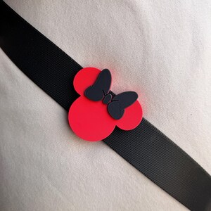 May include: A red and black Minnie Mouse bow-shaped accessory is attached to a black strap. The bow has a red base with two circular shapes and a black bow in the center. The strap is angled across a beige fabric.