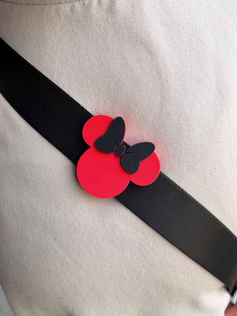 May include: A red and black Minnie Mouse bow-shaped accessory is attached to a black strap. The bow has a red base with two circular shapes and a black bow in the center. The strap is angled across a beige fabric.