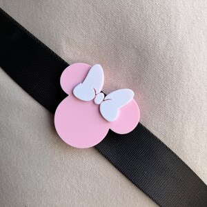 May include: A pink Minnie Mouse bow-shaped accessory with white bow details, set against a black strap and a beige background. The accessory is a playful design, likely for a bag or clothing.