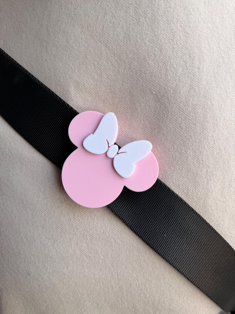 May include: A pink Minnie Mouse bow-shaped accessory with white bow details, set against a black strap and a beige background. The accessory is a playful design, likely for a bag or clothing.