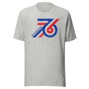 May include: A light gray t-shirt featuring a distressed graphic of the number "76" in red, white, and blue, with the word "Bicentennial" printed diagonally. The shirt has short sleeves and a crew neck.