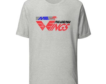 Könnte beinhalten: Hellgraues T-Shirt mit dem Text "MIL Philadelphia WINGS" in Rot, Blau und Grau. Das Design zeigt eine laufende Figur. Das Shirt hat einen Rundhalsausschnitt und kurze Ärmel.