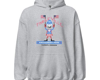 Könnte beinhalten: Hellgrauer Kapuzenpullover mit einem Aufdruck eines Zeichentrickcharakters, der Fahnen mit der Aufschrift "USA" hält. Der Text "THRILLVILLE" wölbt sich über der Figur, darunter "AMUSEMENT PARK" und "TURNER, OREGON".