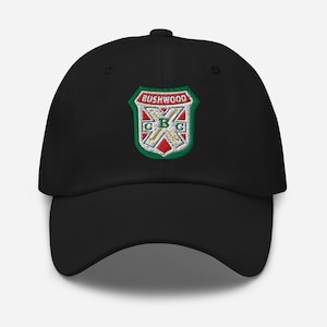 May include: Black baseball cap with a curved brim. The cap features an embroidered crest with the text "BUSHWOOD" above a design with the letters "CBC" in green, red, and white.