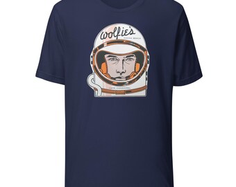 Könnte beinhalten: Marineblaues T-Shirt mit einer Grafik eines Astronautenhelms. Der Text auf dem Helm lautet "wolfie's cocoa beach" und "cape canaveral". Das Gesicht des Astronauten ist sichtbar. Das Design ist in Weiß, Orange und Pfirsich gehalten.