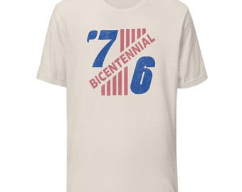 May include: A cream-colored short-sleeve t-shirt featuring a distressed graphic design. The print displays the number 76 in blue with red vertical stripes between the digits and the word BICENTENNIAL written diagonally in red across the center.