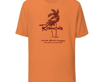 Könnte beinhalten: Orangefarbenes T-Shirt mit einer dunkelroten Grafik von Palmen und dem Text "Ramon's" über "COCOA BEACH FLORIDA" und "THE TOAST OF THE COAST". Das Shirt ist aus einem weichen Material.