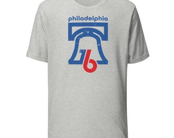 May include: A light gray t-shirt featuring a distressed blue and red graphic of the Liberty Bell and the word "philadelphia" in blue. The graphic is centered on the front of the shirt.