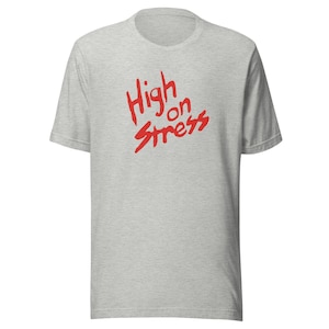 May include: A light grey t-shirt featuring the red text "High on Stress" in a handwritten style across the front. The shirt has short sleeves and a crew neck.