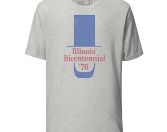 May include: A heather gray short-sleeve t-shirt featuring a blue graphic of a top hat and a curved shape below it. The text "Illinois' Bicentennial '76" is printed in red between the hat and the curved shape. The shirt has a crew neckline.