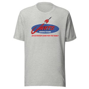 May include: A light grey t-shirt with a vintage-style graphic. The design features the word "Jarts" in red script, a blue oval, and a red and black missile graphic. The text "MISSILE GAME" and "AN OUTDOOR GAME FOR THE FAMILY" are also included.