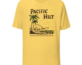 May include: Yellow t-shirt featuring a tropical scene with palm trees, a hut, and the text "PACIFIC HUT". Below the graphic is the text "WESTGATE SHOPPING PARK • BROCKTON, MA". The shirt has short sleeves and a crew neck.