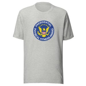 Könnte beinhalten: Ein hellgraues T-Shirt mit einem kreisförmigen Presidential Physical Fitness Award-Emblem. Das Emblem ist blau mit goldenen Sternen, einem gelben Adler und dem Text "Presidential Physical Fitness Award".