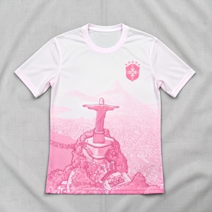 Op de afbeelding: Een t-shirt met korte mouwen in wit- en roze tinten met een grafische print van het Christus de Verlosser-beeld dat uitkijkt over een stadslandschap. Op de borst staat een schildvormig embleem met daarboven vijf roze sterren.