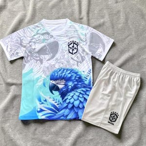 May include: White football kit featuring a blue macaw design on the jersey, with the Brazilian flag emblem. The shorts are white with the Brazilian flag emblem and the word "BRASIL".