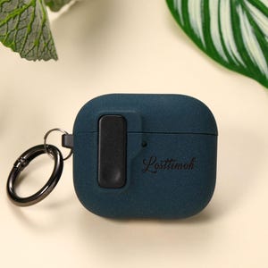 Può includere: Una custodia per AirPod color verde acqua con un fermaglio nero e un pulsante rettangolare nero. La custodia ha la scritta "Losttimoh" stampata sopra. La custodia è su una superficie color crema con foglie verdi sullo sfondo.