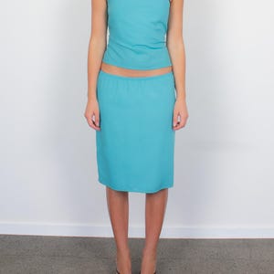 Könnte beinhalten: Ein zweiteiliges türkisfarbenes Outfit. Das Oberteil ist ein One-Shoulder-Design mit braunen Zierleisten. Der Rock hat die passende türkisfarbene Farbe und reicht bis zu den Knien. Das Outfit wird mit schwarzen Riemchensandalen kombiniert.