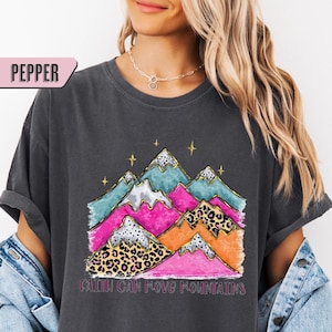 Könnte beinhalten: Dunkelgraues T-Shirt mit einem farbenfrohen Berg-Motiv. Das Design umfasst rosa, türkis, orange und Leopardenmuster-Elemente. Der Text "Faith Can Move Mountains" ist darunter gedruckt. Das T-Shirt hat eine lockere Passform.