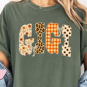 May include: A moss green short-sleeve t-shirt featuring the word GIGI printed in large letters. Each letter has a distinct pattern: floral, leopard print, orange and white gingham, and brown polka dots, accented with small light-colored bows.