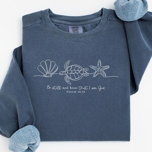 May include: A slate blue long-sleeve sweatshirt featuring a white line art graphic of a seashell, a sea turtle, and a starfish. Below the graphic, the text reads: Be still and know that I am God, PSALM 46:10. The garment has a crew neckline.