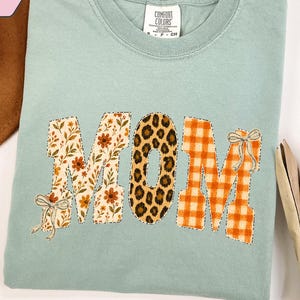 May include: A sage green crewneck t-shirt with the word MOM printed in large block letters. The M letters feature floral and orange gingham patterns with small bows, while the O features a brown and black leopard print pattern.