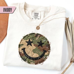 May include: Ivory-colored t-shirt with a smiley face design. The smiley face is a camouflage pattern with black star eyes and a cracked black smile. A gold necklace with a pendant is draped on the shirt.