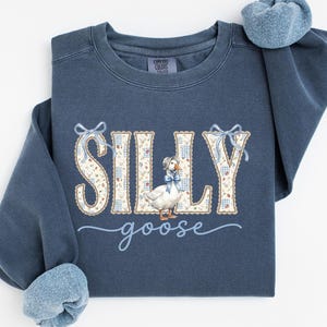 Pode incluir: Uma camisola azul-escura de gola redonda com a palavra SILLY em letras grandes e claras com um padrão floral e de xadrez. Um ganso branco com uma touca e um laço azul está no centro, com a palavra goose escrita por baixo em letra cursiva azul.
