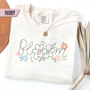 May include: An ivory-coloured t-shirt featuring the word "Bloom" in a playful font, with pastel-coloured line-drawn flowers. A gold necklace with a pendant is draped on the shirt. The shirt is a Comfort Colors brand.