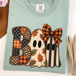 May include: A light green t-shirt featuring a graphic with the word BOO. The letter B is a patchwork of black polka dots, leopard print, and orange plaid. Two ghosts follow, one with autumn leaves and one with black stripes and an orange plaid bow.